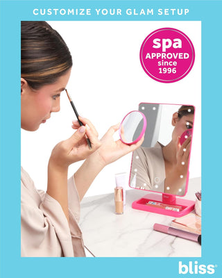 A woman applies makeup using a small brush while looking into an LED Vanity Mirror (with Built-In Speaker & Hands-Free Calling) on a vanity table. Various makeup items are next to her. The text at the top reads, "CUSTOMIZE YOUR GLAM SETUP," and a pink circle states, "spa APPROVED since 1996." The bottom right says, "bliss.