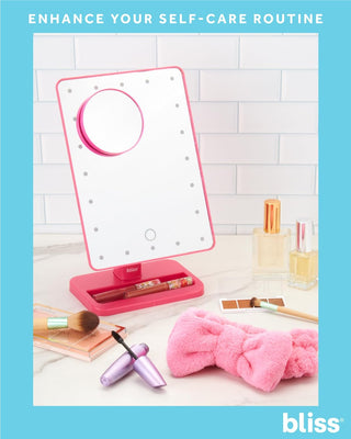 A vanity set featuring a bliss LED Vanity Mirror (with Built-In Speaker & Hands-Free Calling), complemented by an array of beauty products including nail polishes, makeup brushes, an eyeshadow palette, a pink fluffy headband, and curling tools. Text at the top reads, "Enhance Your Self-Care Routine.