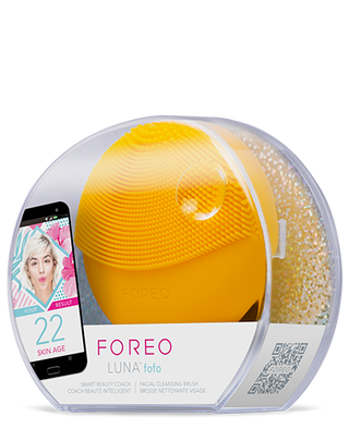 A FOREO LUNA "fofo" Smart Facial Cleansing Device in vibrant yellow, presented in a clear plastic case. The packaging also shows a smartphone displaying a woman's face with "22 Skin Age" written on the screen. The text "Smart Beauty Coach Facial Cleansing Brush" highlights its personalized cleansing routine and skin analysis features below.