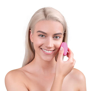 A person with long blonde hair and no visible clothing smiles at the camera while holding a FOREO LUNA "fofo" Smart Facial Cleansing Device to their cheek. The background is white, creating a clean and minimalistic look.