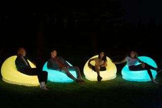 Four people are sitting on Pool Candy's Illuminated LED Color-Changing Chairs made from waterproof PVC vinyl in a dark outdoor setting. The chairs glow in shades of green and blue, providing a colorful ambiance. The group appears to be enjoying a relaxed, casual conversation at night, using the included remote to adjust the lighting as needed.