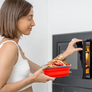 A person with short brown hair wearing a white tank top is placing an Amoré Kitchen Collapsible & Reusable Pizza Storage Container filled with pizza into a black microwave oven. They have a slight smile and are holding the microwave door open with their right hand.