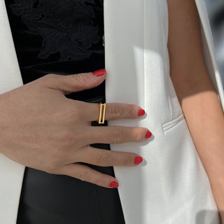 A person wearing a white jacket and black clothing underneath places their hand, adorned with red painted nails and a Sonia Hou Jewelry CEO Double Bar Ring, on their chest.