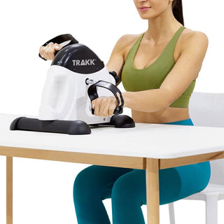 A woman in a green sports bra and blue leggings uses the white and black "Office & Under Desk Exercise Bike with LCD Display" by Trakk on a table. Seated on a chair, she grips the handlebars and pedals with her hands, showcasing its compact design that's perfect for any workspace.