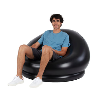 A smiling man with short dark hair sits comfortably in a large, round, Pool Candy 8-Ball Black & White Inflatable Chair. He is wearing a light blue t-shirt, dark shorts, and white sneakers. The background is plain white, highlighting the portable and versatile design of the chair and the individual.