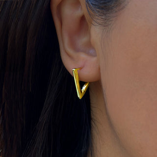 Close-up of a person's ear adorned with the Sonia Hou Jewelry's Achieve Triangle Huggie Earrings. These sleek, gold earrings exhibit a modern design with their downward-hanging triangular shape, serving as a motivational reminder to embrace change. The background brings out the contrast of the person’s dark hair and skin.