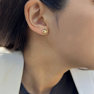 Close-up of an ear adorned with a small, gold Evil Eye Stud Earring from Sonia Hou Jewelry. The person, embracing cultural traditions, has dark hair pulled back and is wearing a light-colored top.