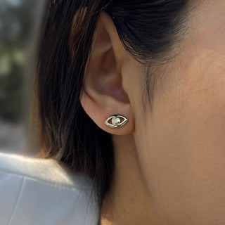 Close-up of a person's ear adorned with Sonia Hou Jewelry's Evil Eye Stud Earrings, featuring a gold-colored eye shape with a silver teardrop at its center. Drawing inspiration from cultural traditions, this unique piece echoes the protective essence of the evil eye symbol. The softly blurred background highlights the intricate jewelry.