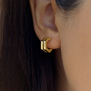 The image shows a close-up of a person's ear wearing Sonia Hou Jewelry's Futuristic Double Hexagon Chubby Huggies. The shiny, gold earrings are positioned on the earlobe, with the person's dark hair partially visible, giving off a sleek and modern vibe.