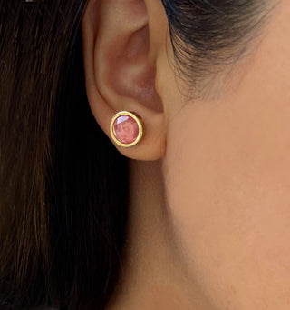 A close-up shot of a person's ear adorned with the Fire Pink Coral Stud Earrings from Sonia Hou Jewelry. The earring features a Pink Coral gemstone encased in a gold setting. The individual has straight dark hair tucked behind the ear, emphasizing the earring.