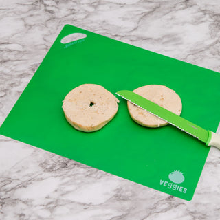 A sliced bagel is placed on a green cutting board from Gia's Kitchen's Flexible Non-Slip Cutting Mats (4-Pack), labeled "VEGGIES" in white text, with a white and green serrated knife resting diagonally across both halves. The non-slip cutting mat is on a marbled surface, providing stability while you slice.