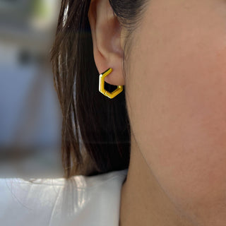 Close-up of a person’s ear wearing Sonia Hou Jewelry's Innovate Hexagon Mini Hoop Earrings in gold. The background is blurred, focusing on the earring's distinct modern style and the person’s short, dark hair. The person is wearing a white top.