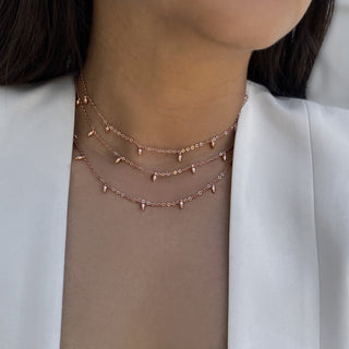 A close-up shows a person wearing the Sonia Hou Jewelry Minimalist Rice Bead Chain Necklace, featuring small, evenly spaced triangular pendants in rose gold. Exuding an aura of wealth, they are also sporting a light-colored blazer, with their hair partially visible on the left side. The background is softly blurred.