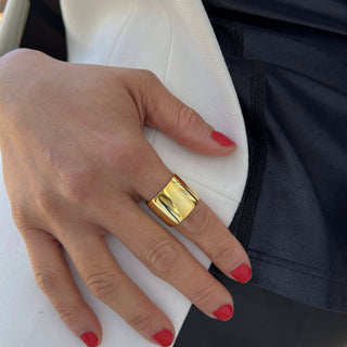 A hand with red painted nails is shown resting on a white surface, making a grand statement. The middle finger is adorned with the Rich Chunky Wide Cigar Band Ring from Sonia Hou Jewelry. The background includes part of a black garment, adding to this wardrobe essential look.