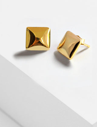 A pair of shiny, gold Square Stud Earrings from Sonia Hou Jewelry are displayed on a minimalistic, light-colored surface. The reflective finish highlights their three-dimensional design, symbolizing strength and unity in communities of color.