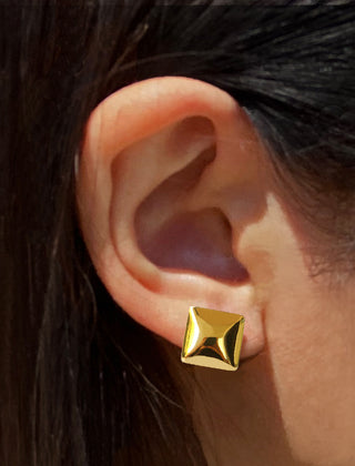 A close-up image captures an ear adorned with a stylish Square Stud Earring by Sonia Hou Jewelry. The gold-colored stud boasts a smooth, shiny surface and is securely fastened to the earlobe. Partially visible dark hair symbolizes resilience and elegance within communities of color.