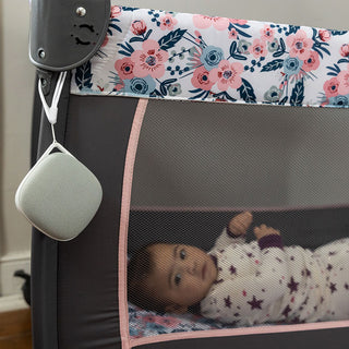 A baby in floral pajamas lies in a crib with a floral-patterned border and mesh sides. A light gray, square-shaped Travelcube Portable Sound Machine from Yogasleep is attached to the corner of the crib. The floor beneath the crib and part of the wall are visible in the background.