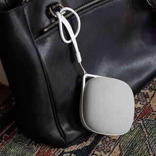 A small, portable sound machine, the Travelcube Portable Sound Machine by Yogasleep, with a white strap hangs from a black leather bag. The speaker has a smooth, rounded design with a light gray fabric covering. The bag features a visible zippered pocket and the scene is set against a patterned fabric background.