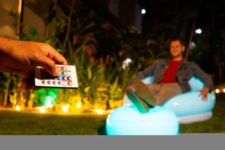 A person uses the remote included with the Pool Candy Illuminated LED Color-Changing Chair to adjust the lighting on an inflatable chair outside at night. In the background, another person relaxes on the vibrant, color-changing chair with their legs crossed. The area is decorated with string lights and foliage.