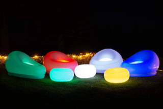 A nighttime garden scene features a variety of Pool Candy Illuminated LED Color-Changing Chairs, equipped with remote controls, and tables arranged in a semi-circle on the grass. Made from waterproof PVC vinyl, the lights display shades of green, red, blue, white, and yellow, creating a vibrant and inviting atmosphere.