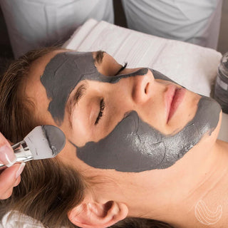 A person lies on a spa bed with their eyes closed while another hand applies INNATUS's "Skin Refresh" Dead Sea Mud Face Mask to their face using a brush. The environment, a serene skincare facility, enhances the mask's detoxification and purification properties for optimal relaxation and skin rejuvenation.